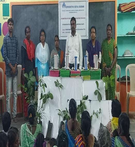 Adult literacy learners seated in front of a blackboard, with slates and potted medicinal plants set out on a table