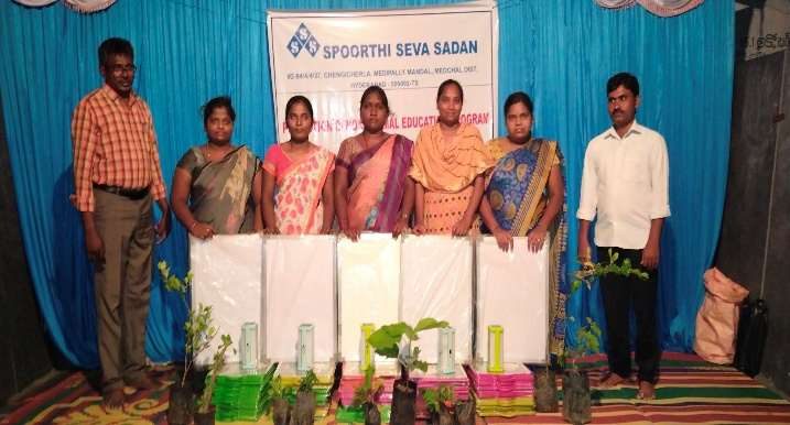 Volunteers holding white boards in front of a Spoorthi Seva Sadan banner, with potted plants at their feet